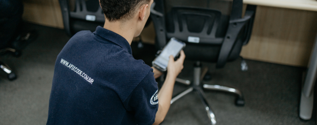 homem de costas fazendo o inventário patrimonial segurando um celular coletando dados mirando em uma cadeira com etiqueta de patrimonio