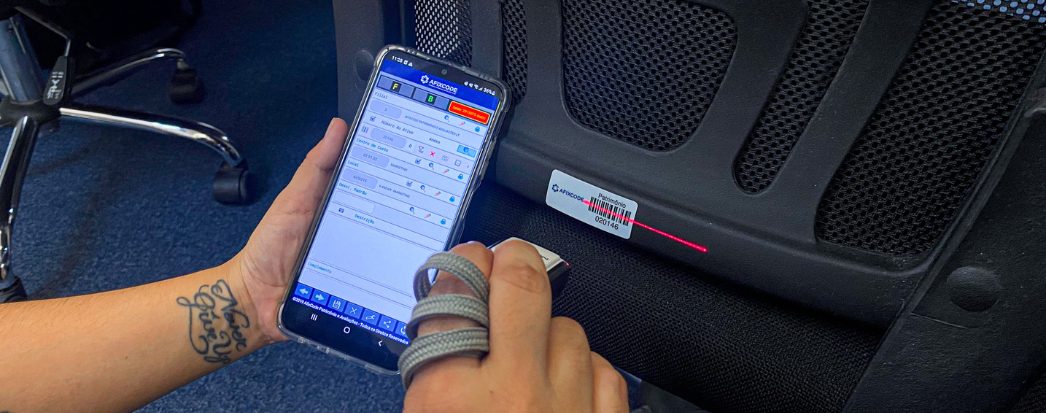 mãos feminina segurando um celular dentro do app afixinv para realizar o inventário, na outra mão está segurando um leitor e apontando para a etiqueta que está colada em um bem